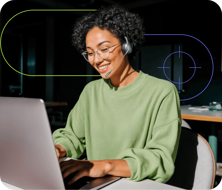 Woman in a headset smiling as she works at a laptop in a dim office