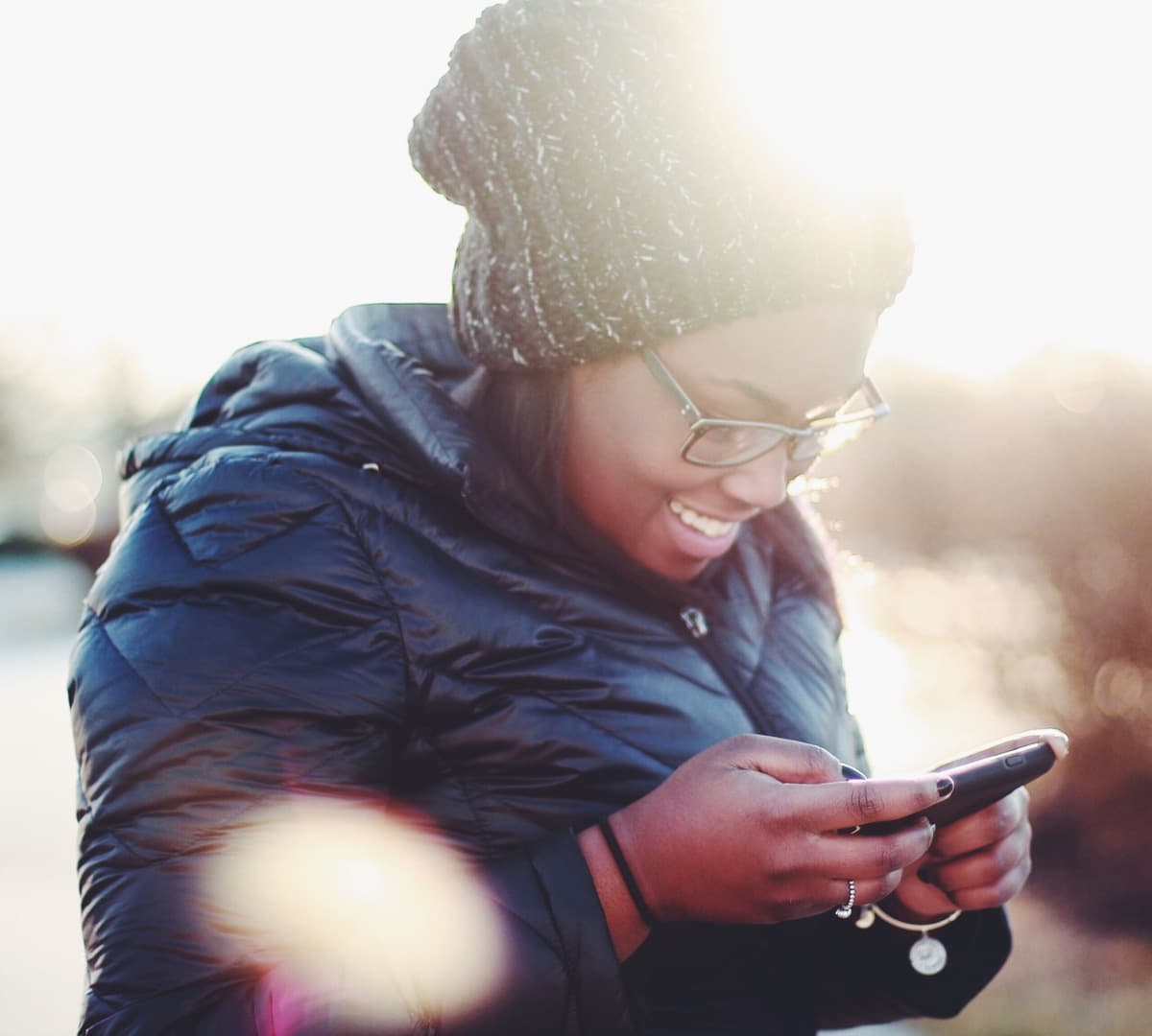 Image of a woman in a winter coat and hat looking down at a mobile phone and smiling.