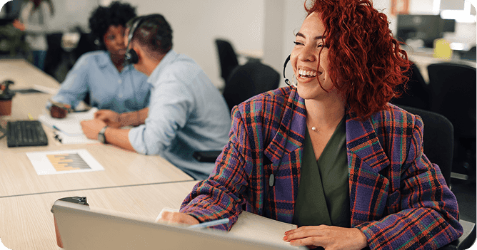 A cheerful customer support representative working at a desk, with colleagues in the background wearing headsets.
