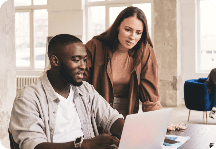 Two colleagues collaborating at a table; one works on a laptop while the other leans in to assist, set in an office space.