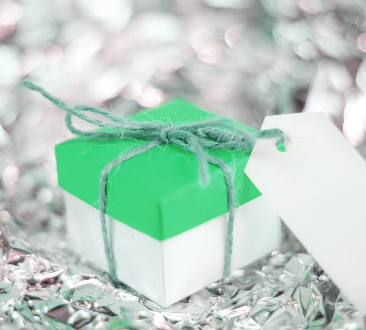 A green and white gift box that has a blank label tied to it with twine. Gift box sits on a silver, reflective surface.